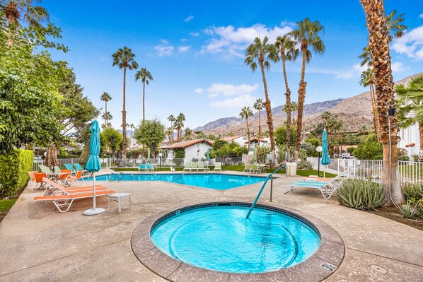 A swimming pool area with lounge chairs and umbrellas, surrounded by palm trees and a mountain backdrop.