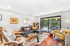 A modern living room with a sectional sofa, a glass coffee table, and a colorful rug. There is a dining area with a table and chairs, and a large window offering a view of greenery outside.