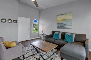 A living room with a gray sofa, a gray armchair, a wooden coffee table, and a patterned rug. There is a door with a window, a small table with a lamp, and wall art.