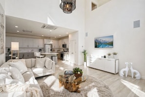 A modern living room with a glass coffee table, a sofa, and a dining area with a white cabinet and plants.
