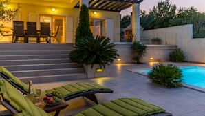 A poolside area with lounge chairs, a table with a bottle and fruit, and a staircase leading to a building.