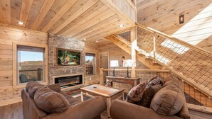 L’intérieur chaleureux d’un chalet avec des murs en bois, une cheminée en pierre, un téléviseur et un escalier.