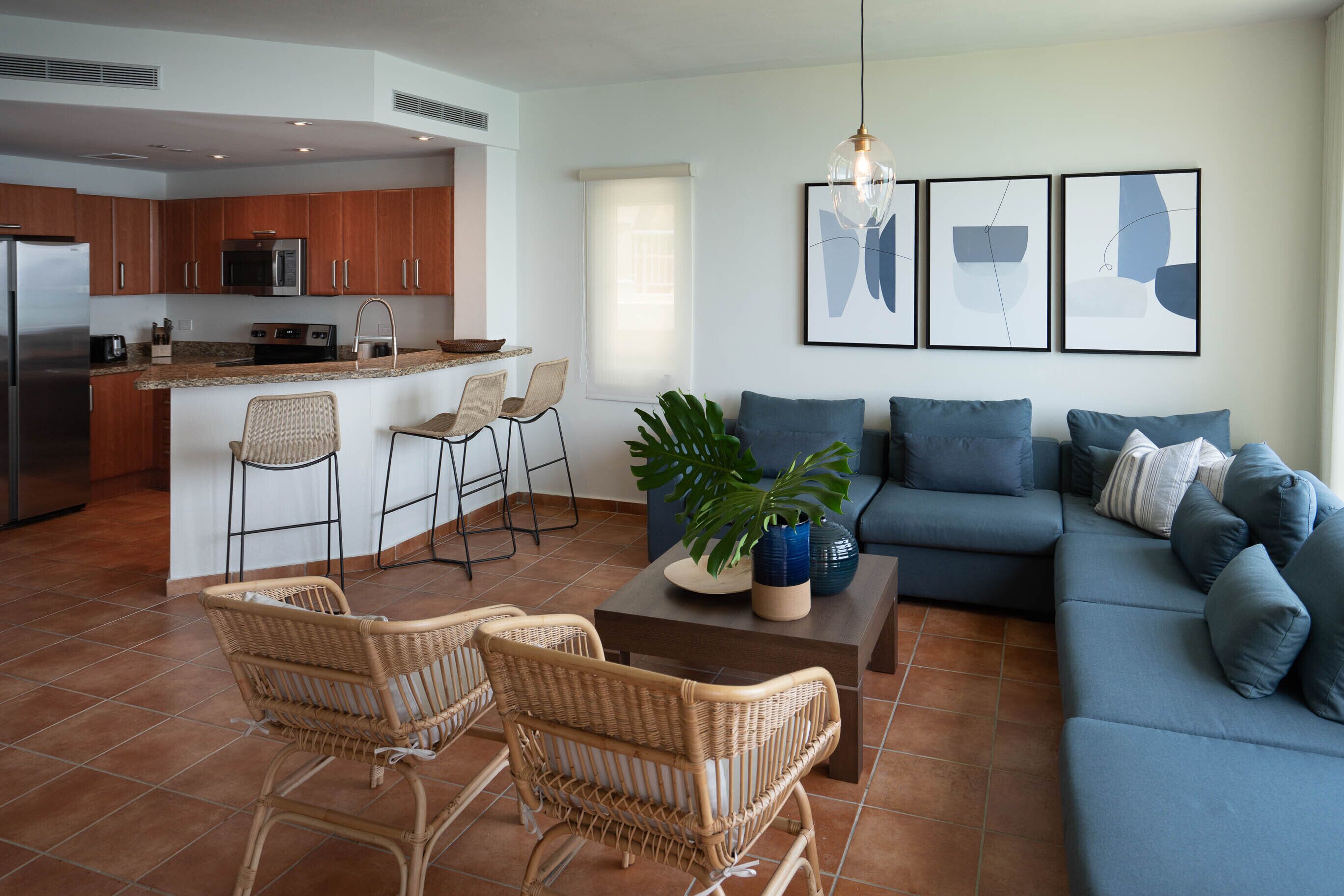 Une cuisine moderne avec un îlot central, un coin repas avec des chaises en osier, un canapé bleu agrémenté de coussins et des œuvres d’art abstraites accrochées au mur.