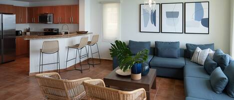 Une cuisine moderne avec un îlot central, un coin repas avec des chaises en osier, un canapé bleu agrémenté de coussins et des œuvres d’art abstraites accrochées au mur.
