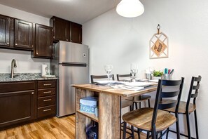 A modern kitchen with dark wooden cabinets, a stainless steel refrigerator, a wooden dining table with chairs, and a pendant light.