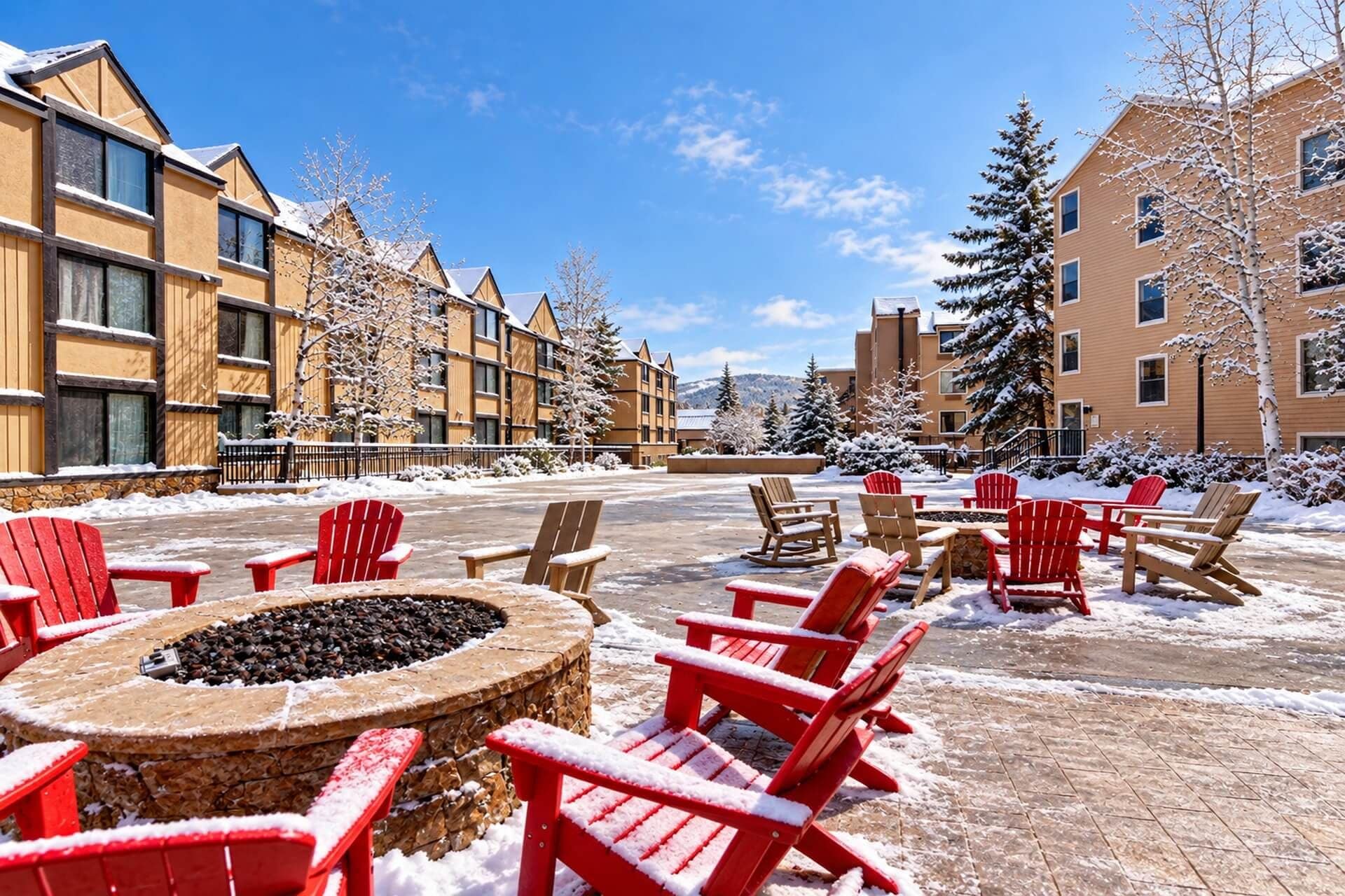 A snow-covered outdoor seating area with red Adirondack chairs and a stone fire pit.