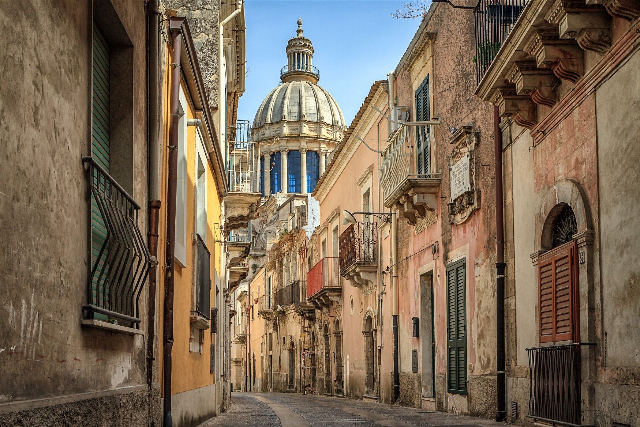 A narrow street with historic buildings, a prominent dome structure in the background, and a mix of balconies and shutters.