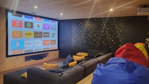 Una sala de estar moderna con una pantalla grande que muestra servicios de streaming, un sofá acogedor, sillones de peluche y una decoración de pared con motivos de estrellas.