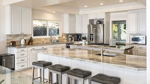 A modern kitchen with white cabinetry, a large island with bar stools, stainless steel appliances, and a view of the outdoors. - Dolphin Manor: Stunning Oceanfront Home w/Pool, Spa, A/C, Private Lanai (Kailua Kona)