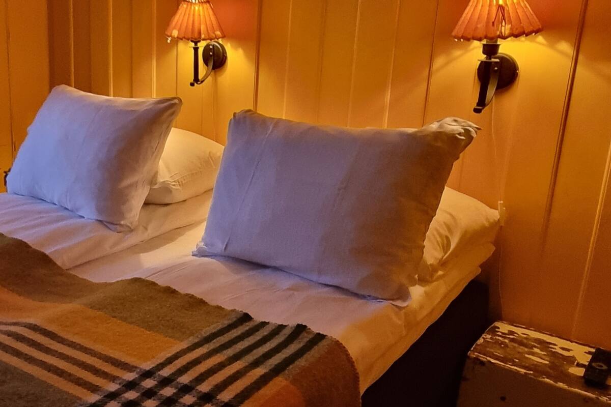 Una cama con ropa de cama blanca, un plaid a rayas y dos lámparas fijadas a la pared.