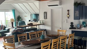 A dining area with a wooden table and chairs, a television in the background, and a kitchen area with cabinets and appliances.