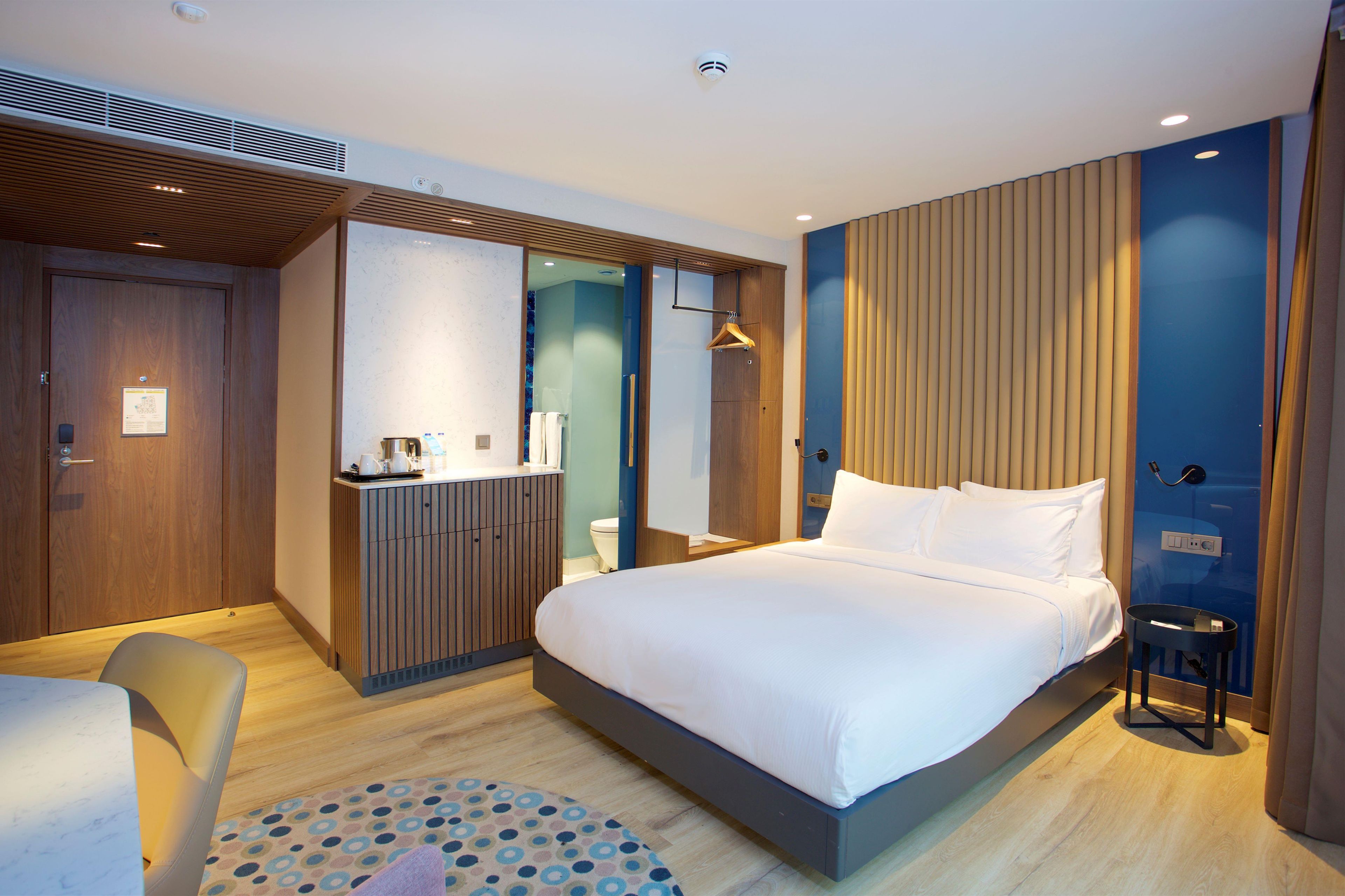 A modern hotel room with a large bed, a wooden wardrobe, a desk with a chair, and a small round table.
