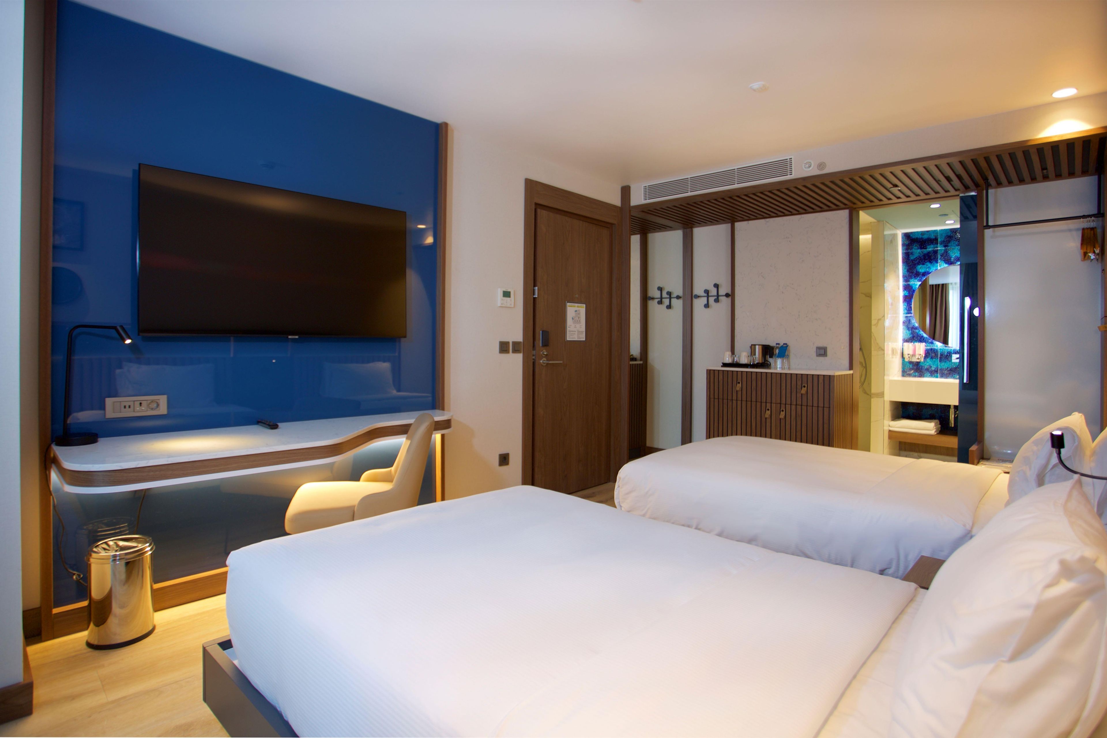 A hotel room with a large flat-screen TV mounted on the wall, a minibar, a wooden door, and a bathroom visible through a glass partition.