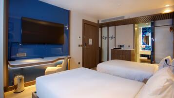 A hotel room with a large flat-screen TV mounted on the wall, a minibar, a wooden door, and a bathroom visible through a glass partition.