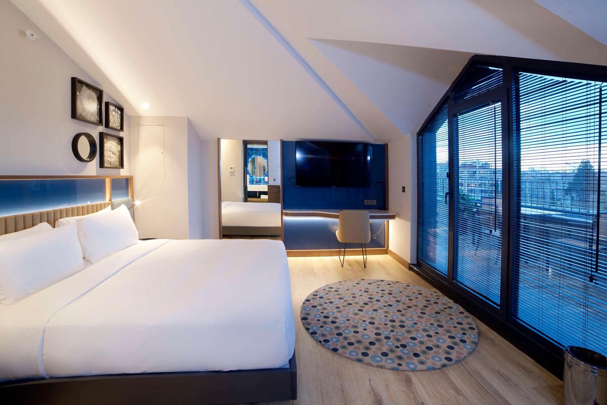 A modern hotel room with a large bed, a desk, a TV, and a large window with blinds.