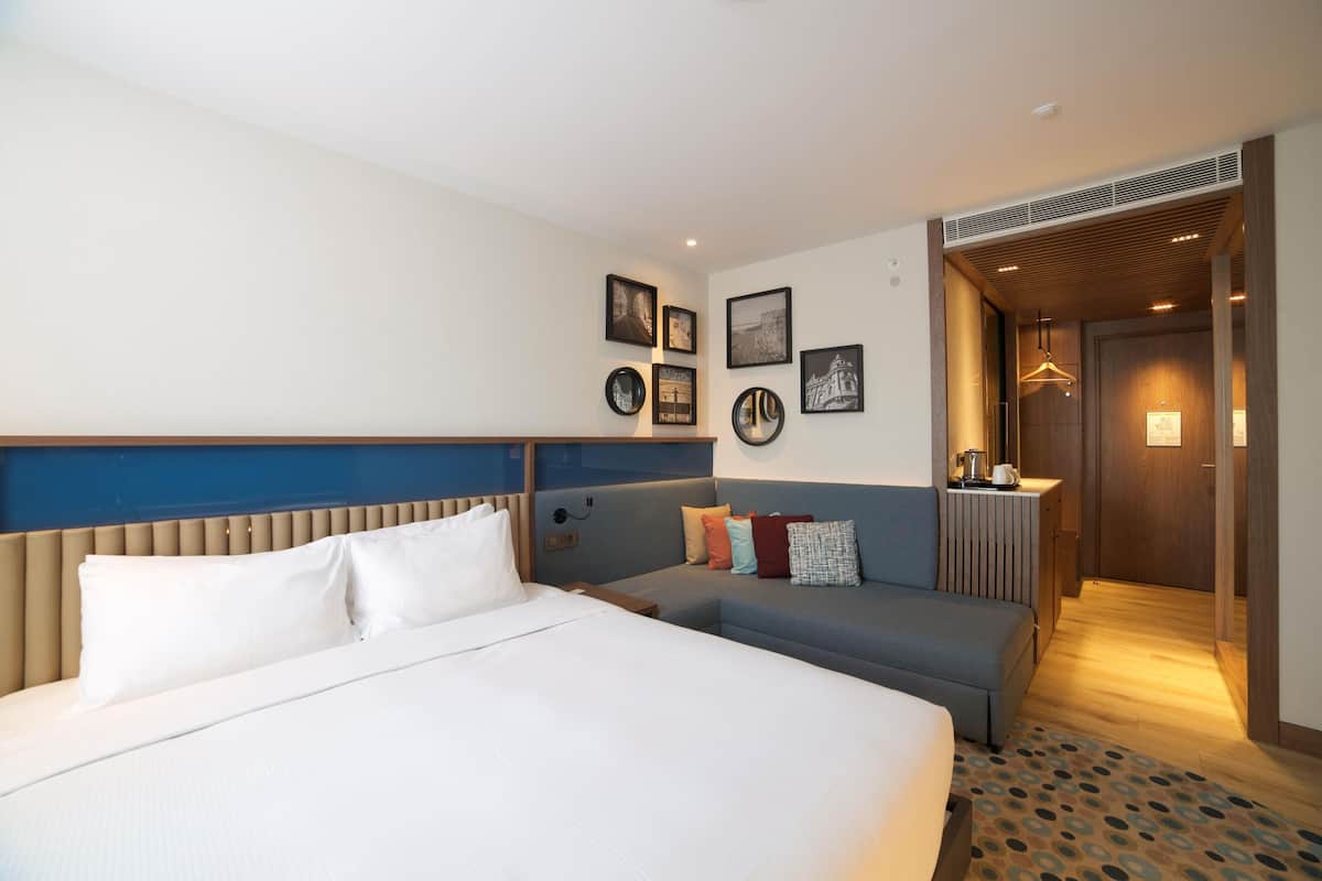 A hotel room with a large bed, a sofa, a desk with a lamp, and a hallway leading to a bathroom.