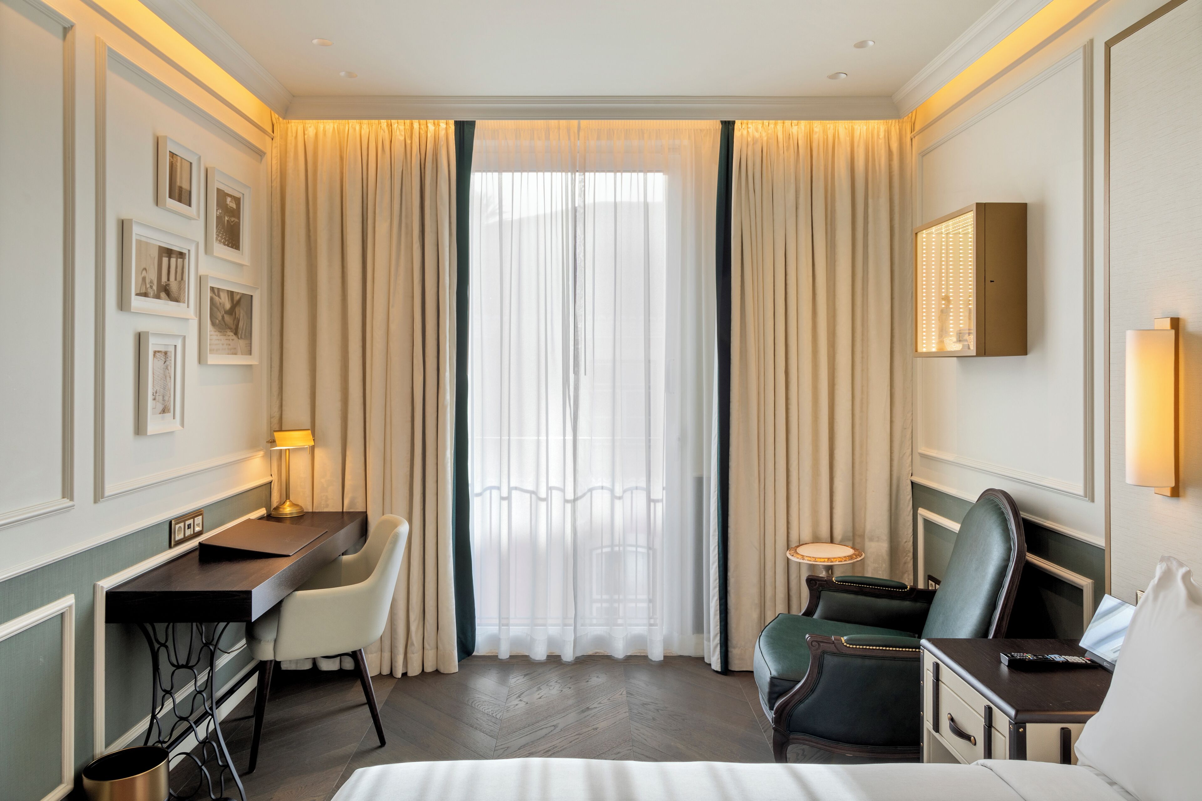 A hotel room with a bed, desk, chairs, a small table, a wall-mounted TV, and a window with curtains.