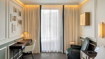 A hotel room with a bed, desk, chairs, a small table, a wall-mounted TV, and a window with curtains.
