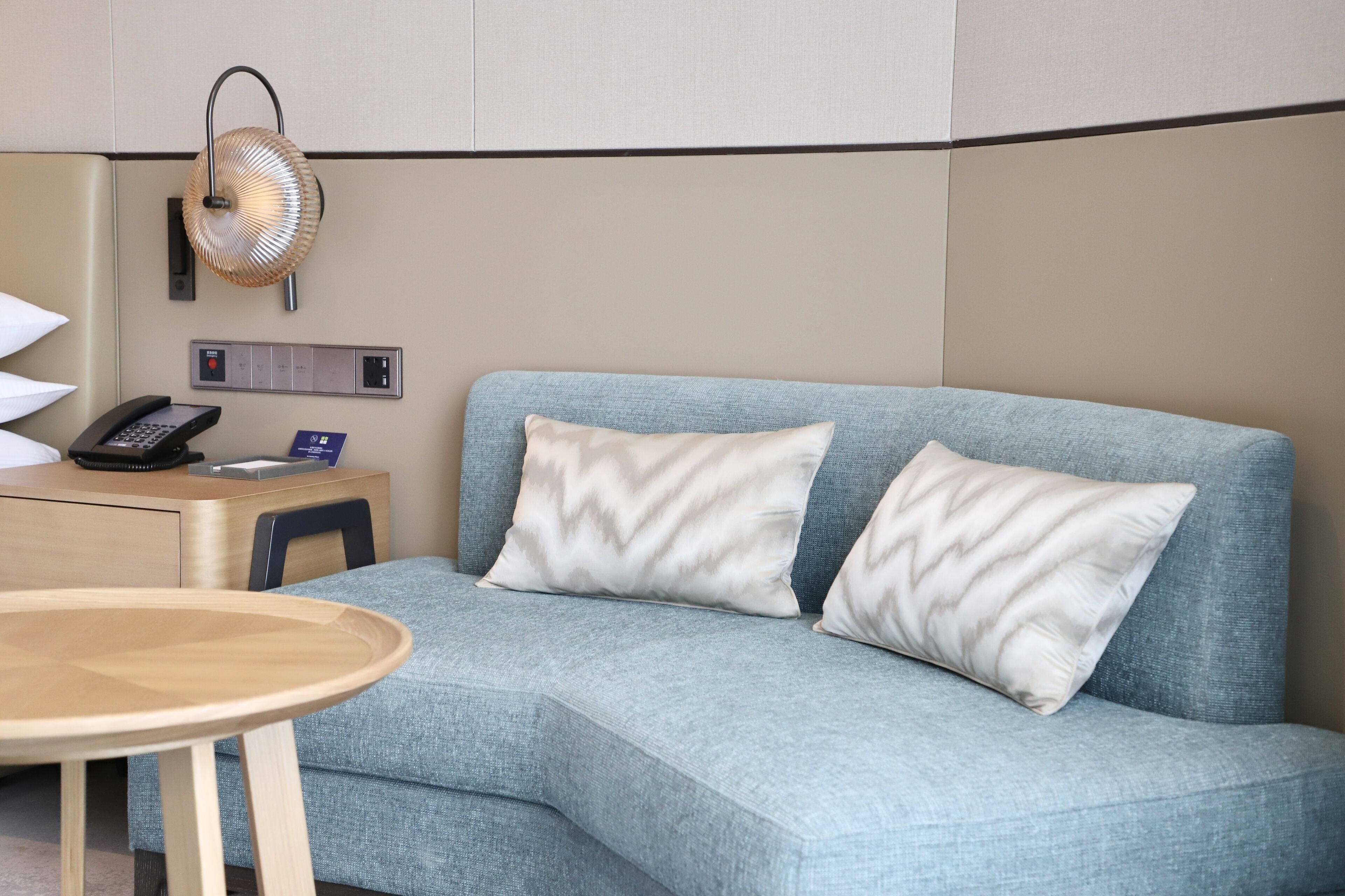 A hotel room with a light blue sofa, two white pillows, a wooden side table, a lamp, and a control panel on the wall.