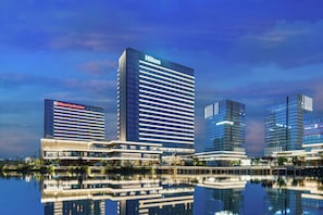 Un complexe hôtelier moderne composé de plusieurs immeubles de grande hauteur, dont un hôtel Hilton, qui se reflète dans une étendue d’eau calme.