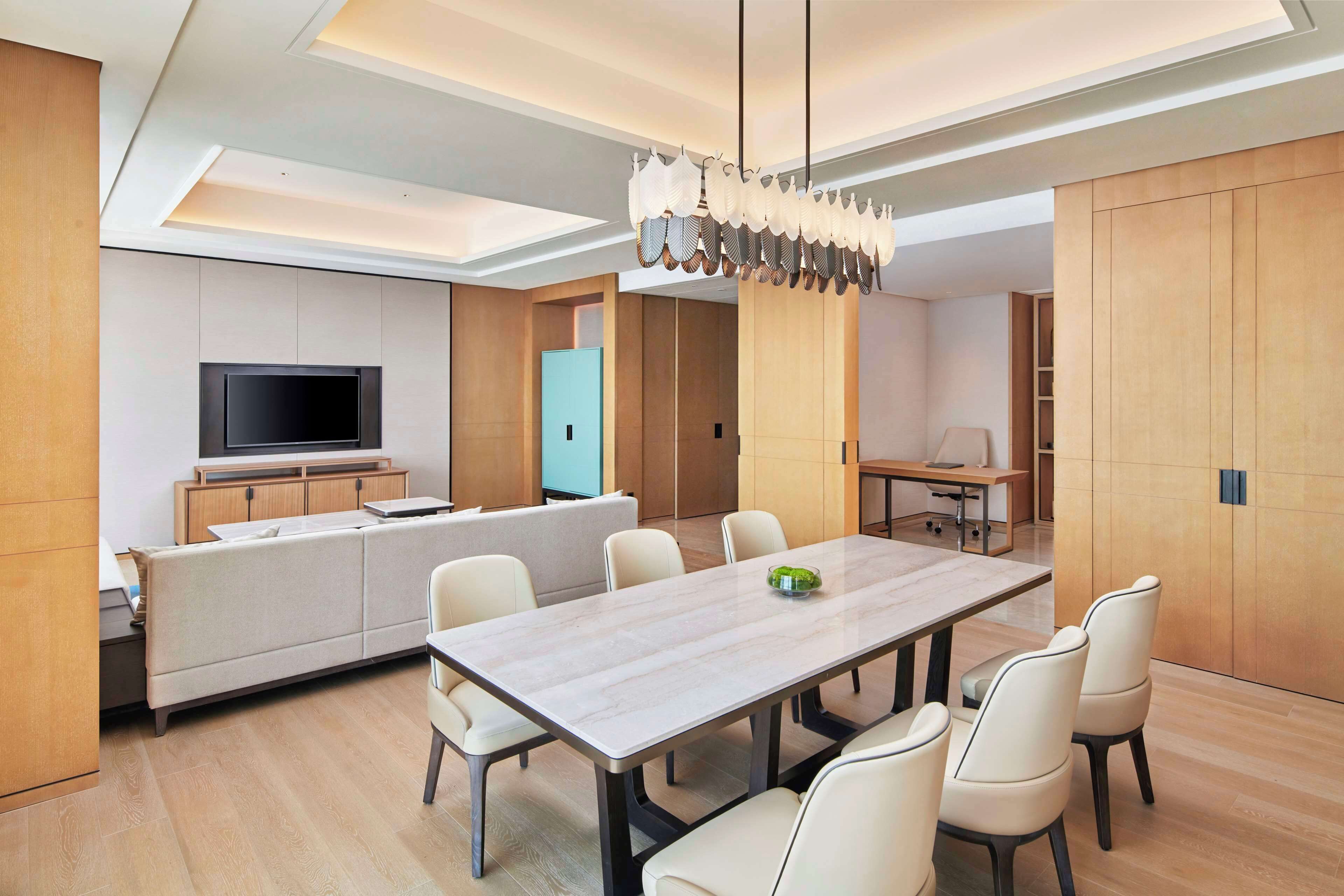 Un espace repas moderne avec une longue table et six chaises, un téléviseur posé sur une étagère intégrée, et un coin cuisine doté de meubles en bois et d’une porte bleue.