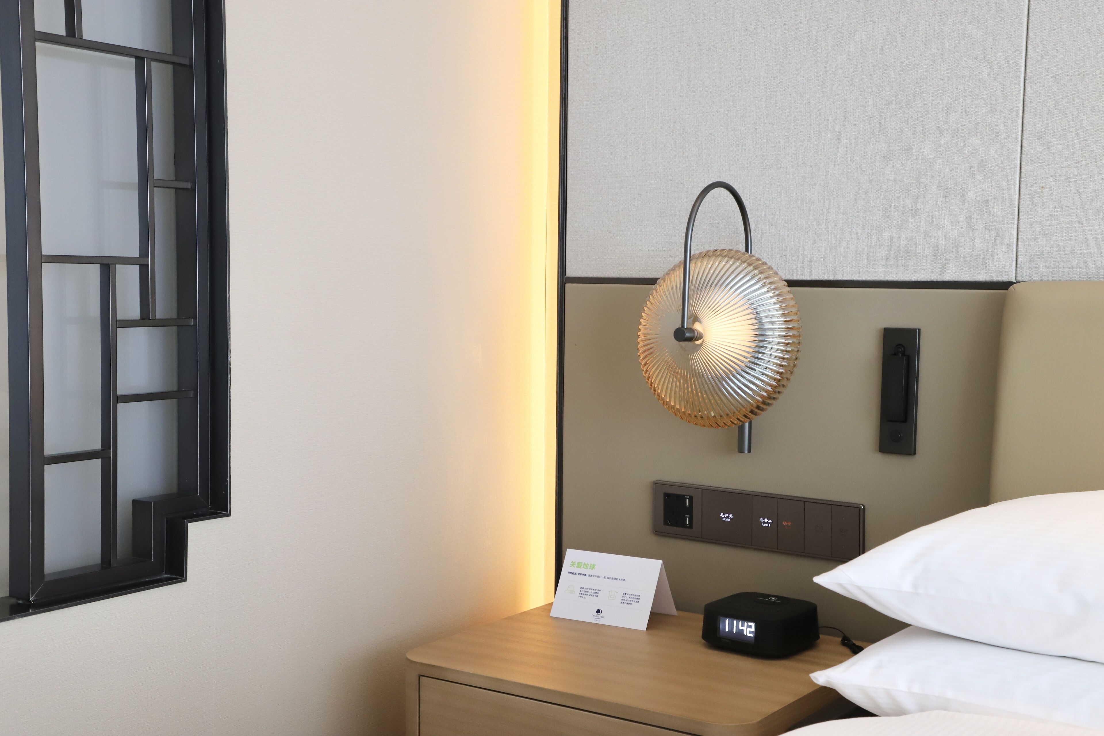 Habitación de hotel ordenada con una mesita de noche, un ventilador de pared, un reloj digital y un cartel que indica los servicios de la habitación.