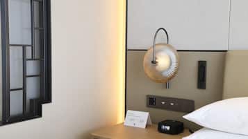 Habitación de hotel ordenada con una mesita de noche, un ventilador de pared, un reloj digital y un cartel que indica los servicios de la habitación.