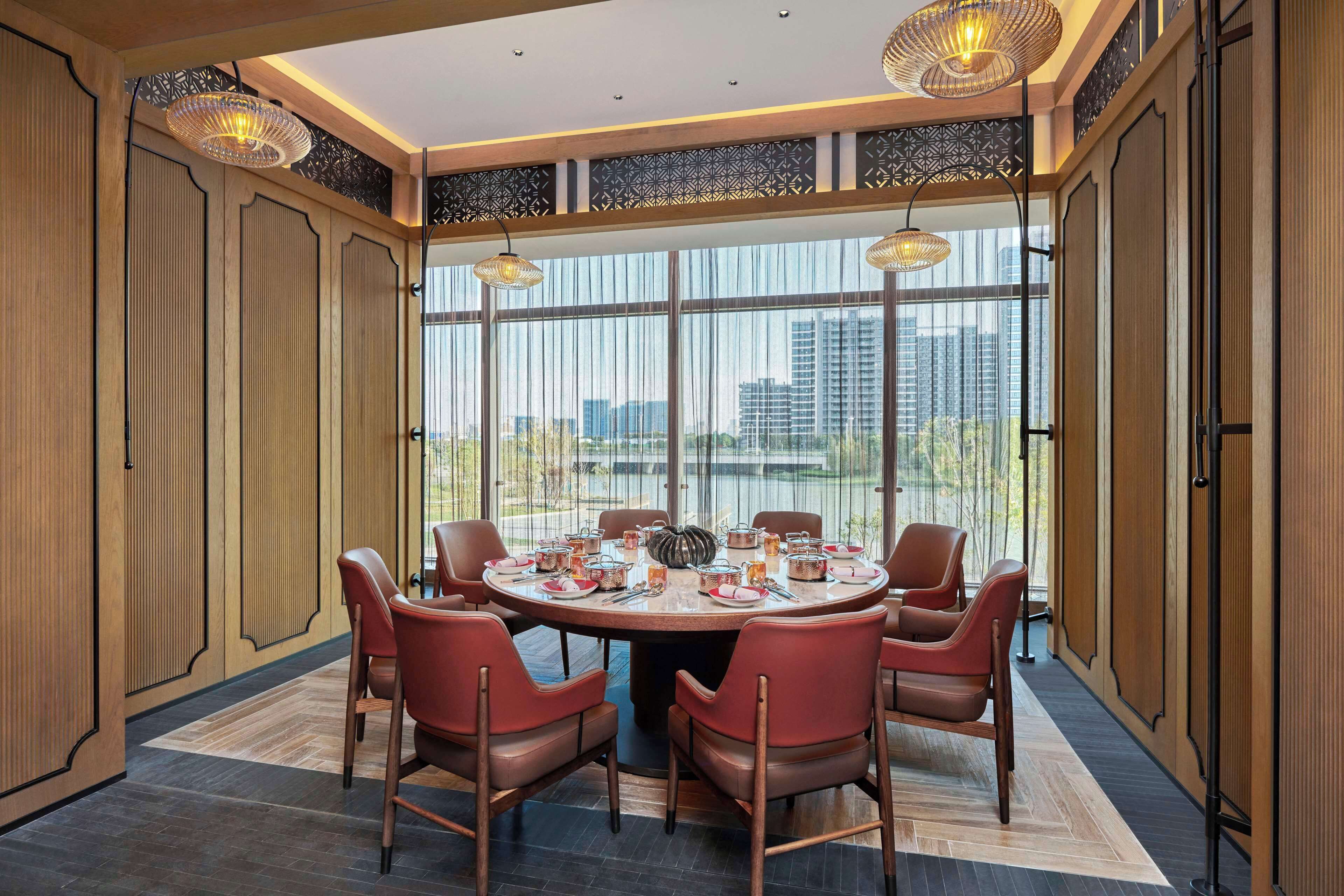 A round dining table with six chairs, set for a meal, in a room with large windows and a view of buildings.