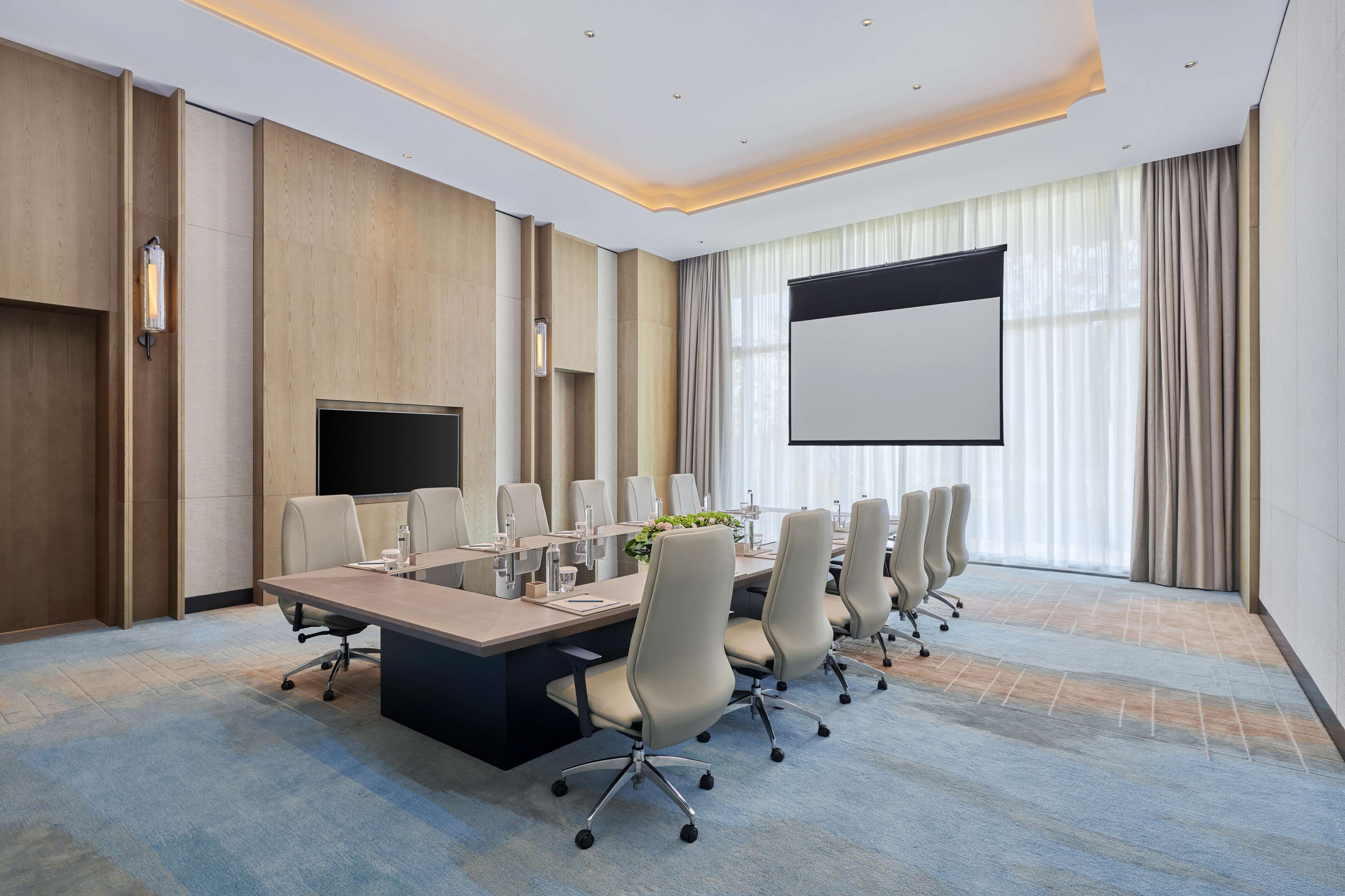 A conference room with a long table, multiple chairs, a projector screen, and a large window with curtains.