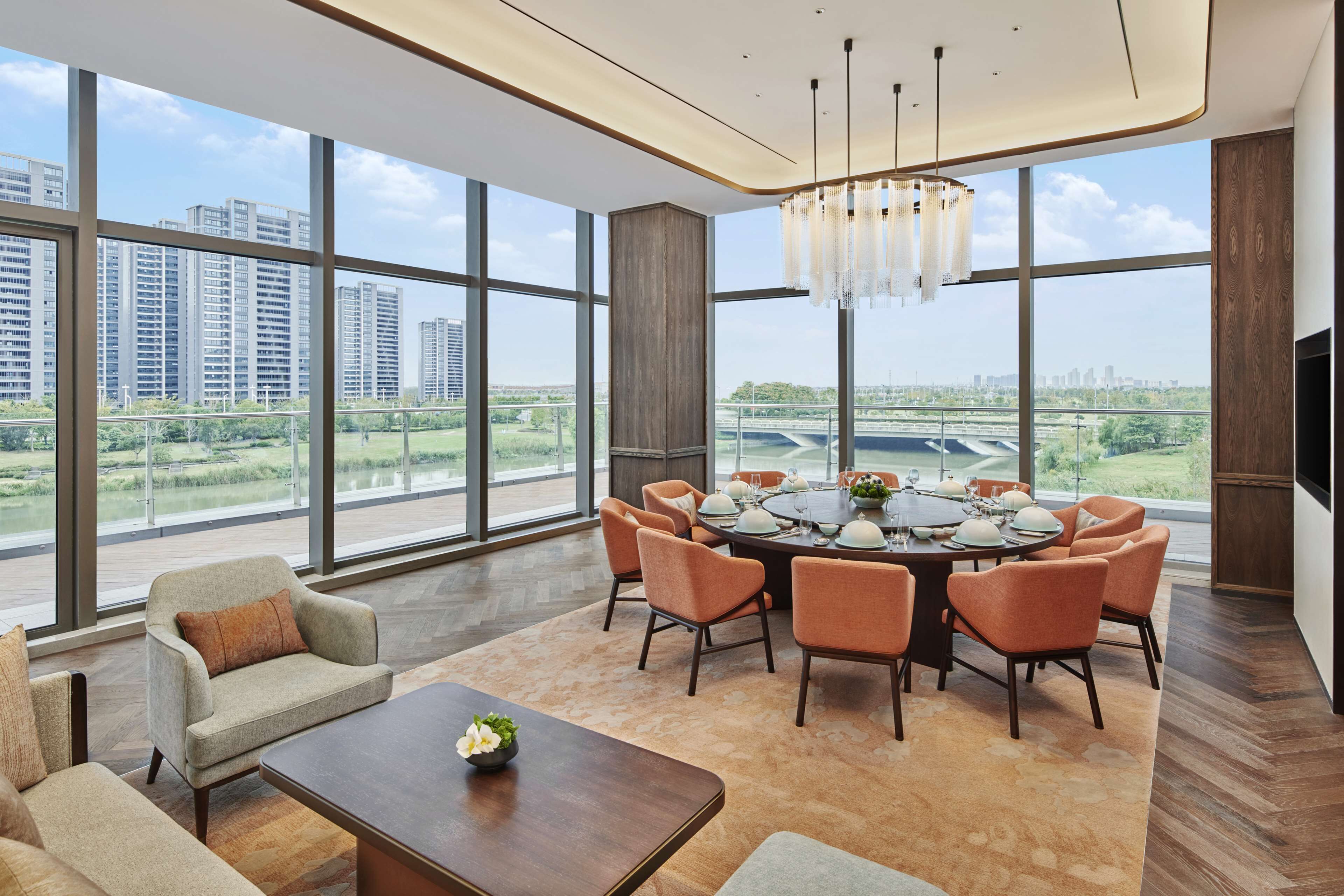 A spacious living room with a large dining table, comfortable seating, and a view of a cityscape through large windows.