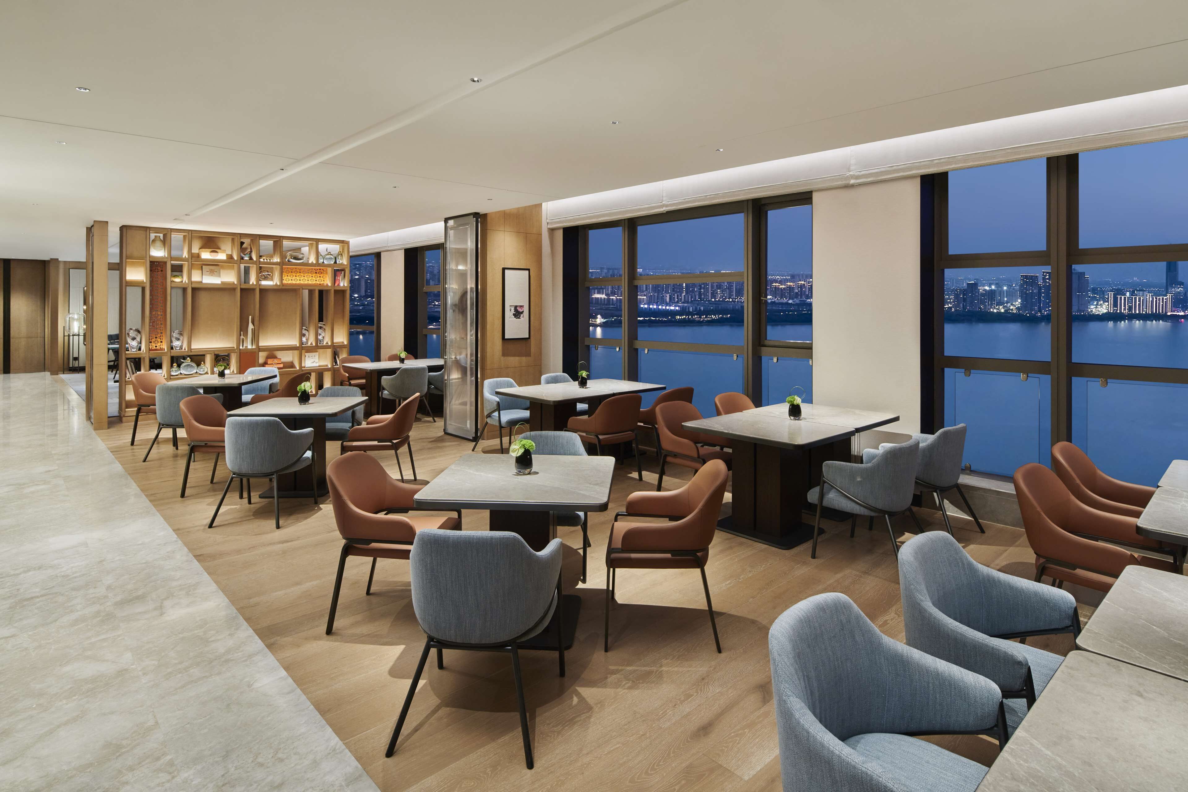 A modern dining area with tables and chairs, large windows with a city view, and a decorative wooden partition with lights.