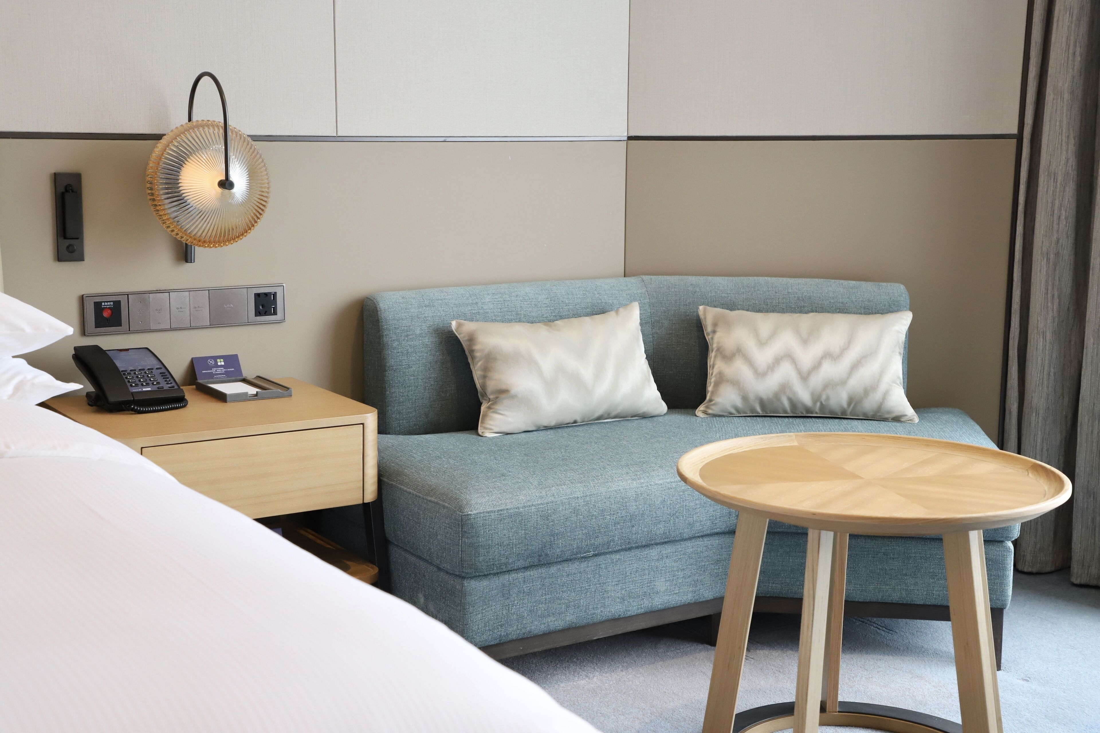 Una habitación de hotel con una cama, un sofá azul con dos almohadas, una mesita de noche con un teléfono y una lámpara, y una pequeña mesa de madera.