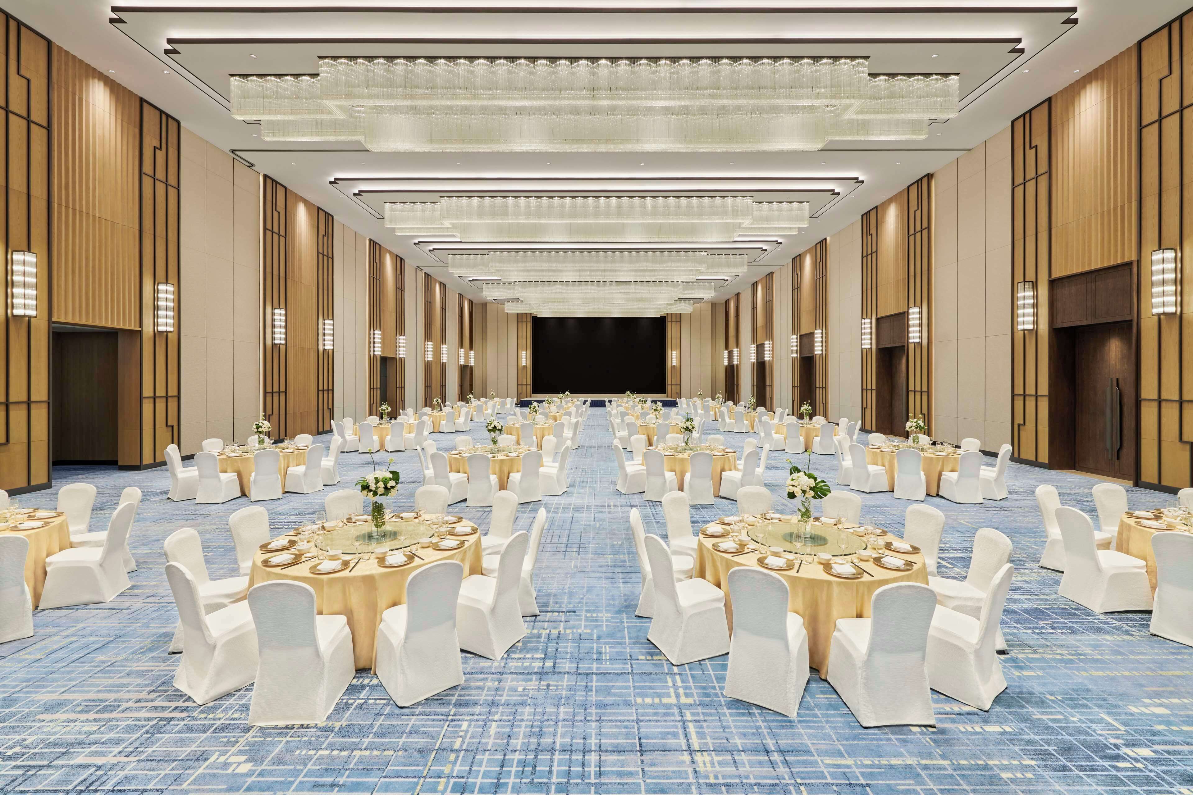A large banquet hall with round tables and white chairs, a blue carpet, and a long table at the front.