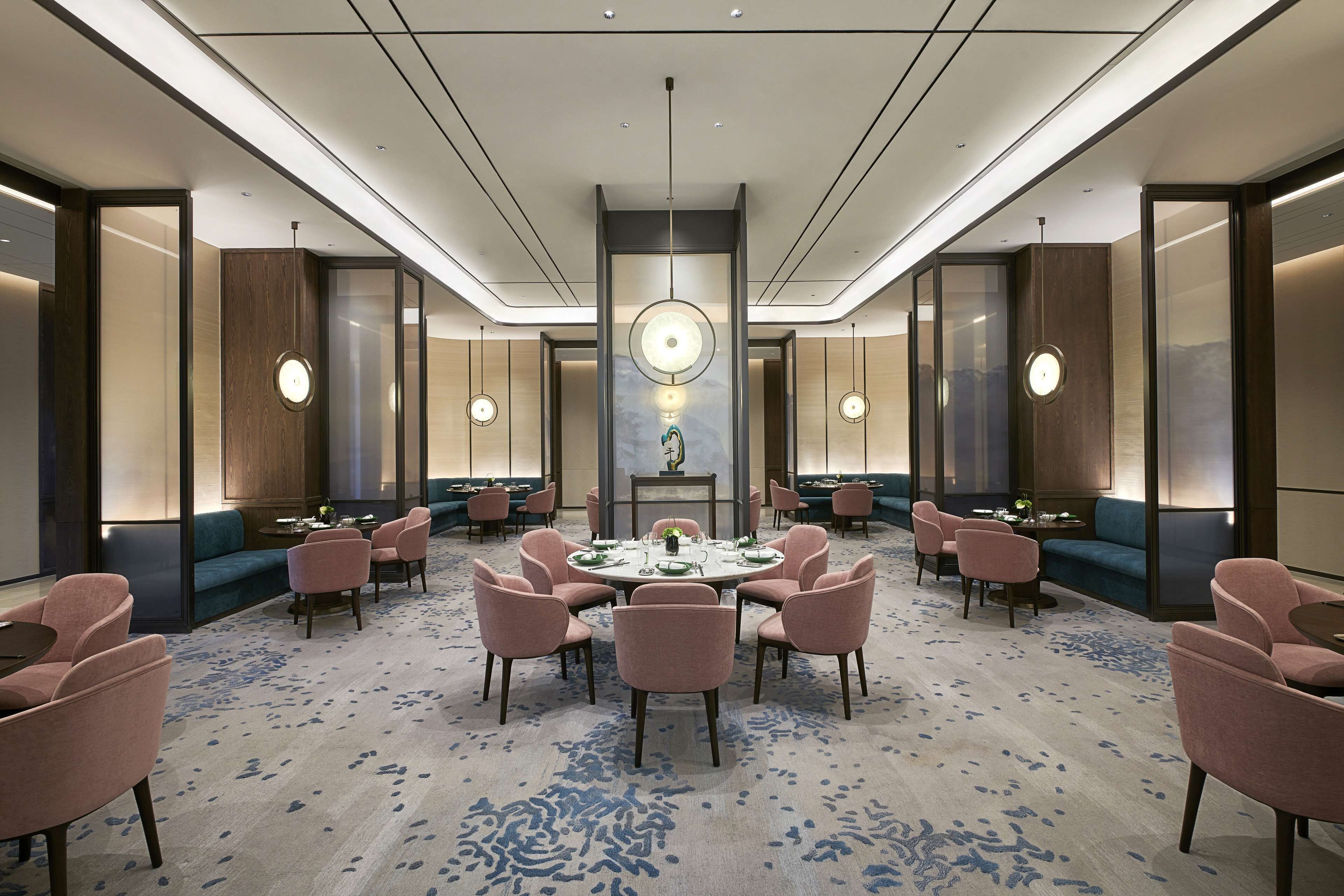 A spacious hotel lobby with a central round table surrounded by chairs, a large chandelier, and a blue patterned carpet.
