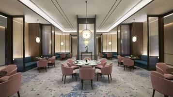 A spacious hotel lobby with a central round table surrounded by chairs, a large chandelier, and a blue patterned carpet.