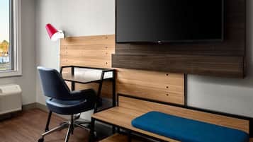 A modern hotel room with a wooden headboard, a flat-screen TV, a blue chair, a small table, and a bench.