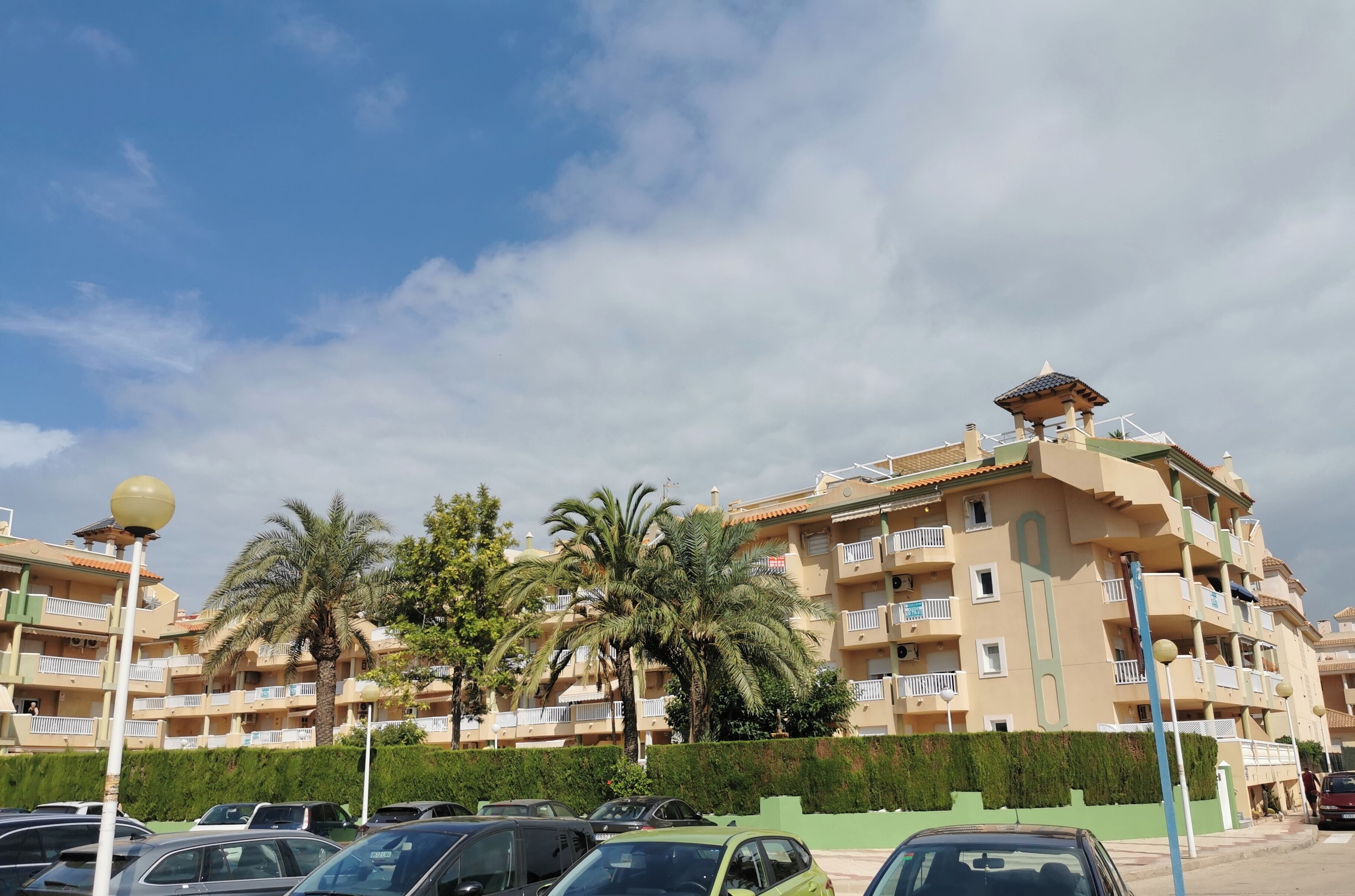 A multi-story residential building with balconies, a parking lot, and palm trees.