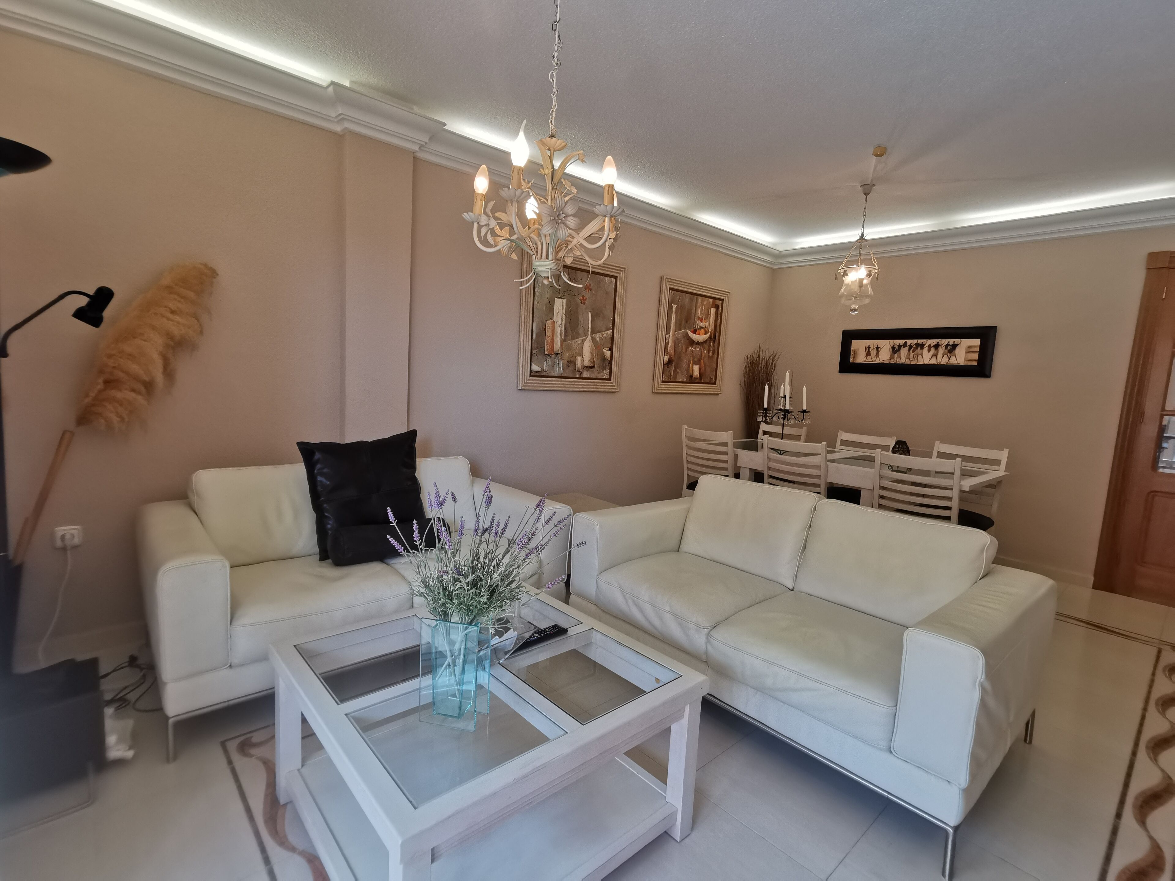 A living room with a white sofa, a coffee table with a vase, a chandelier, and a dining area with chairs and a table.