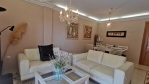 Ein Wohnzimmer mit einem weißen Sofa, einem Couchtisch mit einer Vase, einer Chandelüster und einem Essbereich mit Stühlen und Tisch.