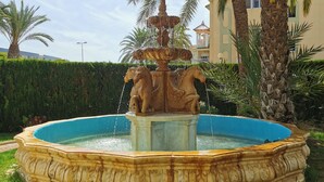 Ein Springbrunnen mit einer zentralen Statue, umgeben von einem kreisförmigen Teich, gelegen in einem angelegten Garten mit Palmen und einer Hecke.