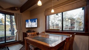 Ein gemütlicher Essbereich mit Holztisch und Stühlen, einem an der Wand befestigten Fernseher und großen Fenstern mit Blick hinaus.