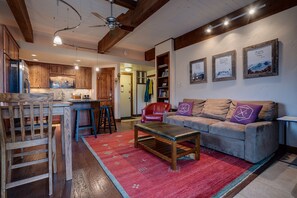 Un salon chaleureux avec un canapé, une table basse et un tapis rouge. On y trouve également un coin cuisine avec des meubles en bois et un tabouret de bar.