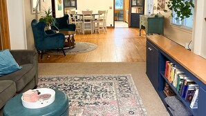 A living room with a sofa, ottoman, and a coffee table with decorative items. There is a dining area with a table and chairs, and a bookshelf filled with books. - Lake home, across the street from a golf course & minutes to shopping and more! (Sauk Centre)