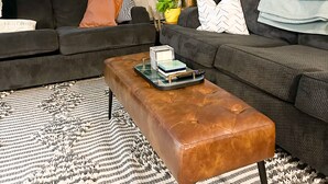 A modern living room with a dark grey sofa, a brown leather ottoman, and a patterned rug. There are two potted plants, a framed artwork on the wall, and a window with a view of trees. - Lake home, across the street from a golf course & minutes to shopping and more! (Sauk Centre)