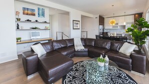 Een moderne woonkamer met een L-vormige bruine leren bank, een glazen salontafel met decoratieve objecten, en een eethoek met een kroonluchter.