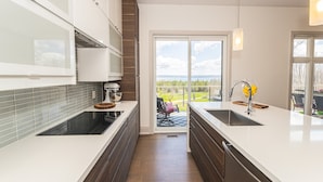 Een moderne keuken met een groot eiland, een ingebouwde oven en uitzicht op de natuur door een glazen deur.