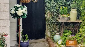Un espace jardin agrémenté de diverses plantes et fleurs en pot, une porte noire et une petite table sur laquelle est posé un vase.