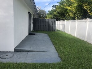 A residential backyard with a concrete pathway leading to a wooden door, surrounded by a white fence and a grassy lawn.