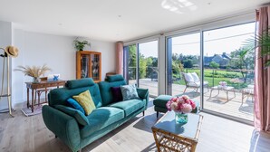 Un salon spacieux avec un grand canapé vert, une table basse en bois et une vue sur un jardin, visible à travers de grandes portes-fenêtres coulissantes.