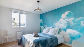 Une chambre avec une grande fenêtre, un lit recouvert d’une couverture bleue, une chaise et un mur peint de nuages.